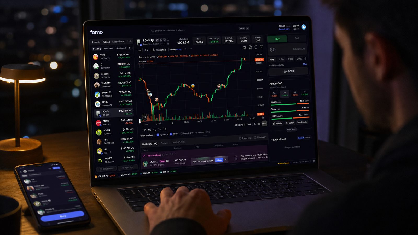 A trader at a desk at night using Fomo Web on a laptop: trending token list, candlestick chart with trader avatars on the candles, and the Buy panel, with the Fomo phone app beside it